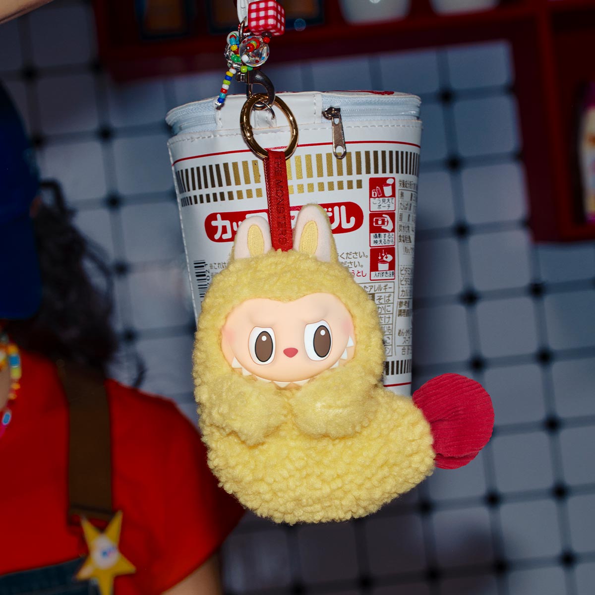 Labubu tempura. Yellow plush keychain with red accents hanging from a cup holder against a grid background