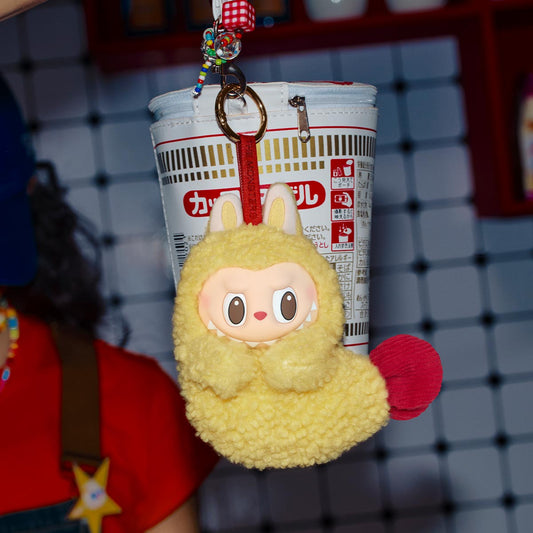Labubu tempura. Yellow plush keychain with red accents hanging from a cup holder against a grid background