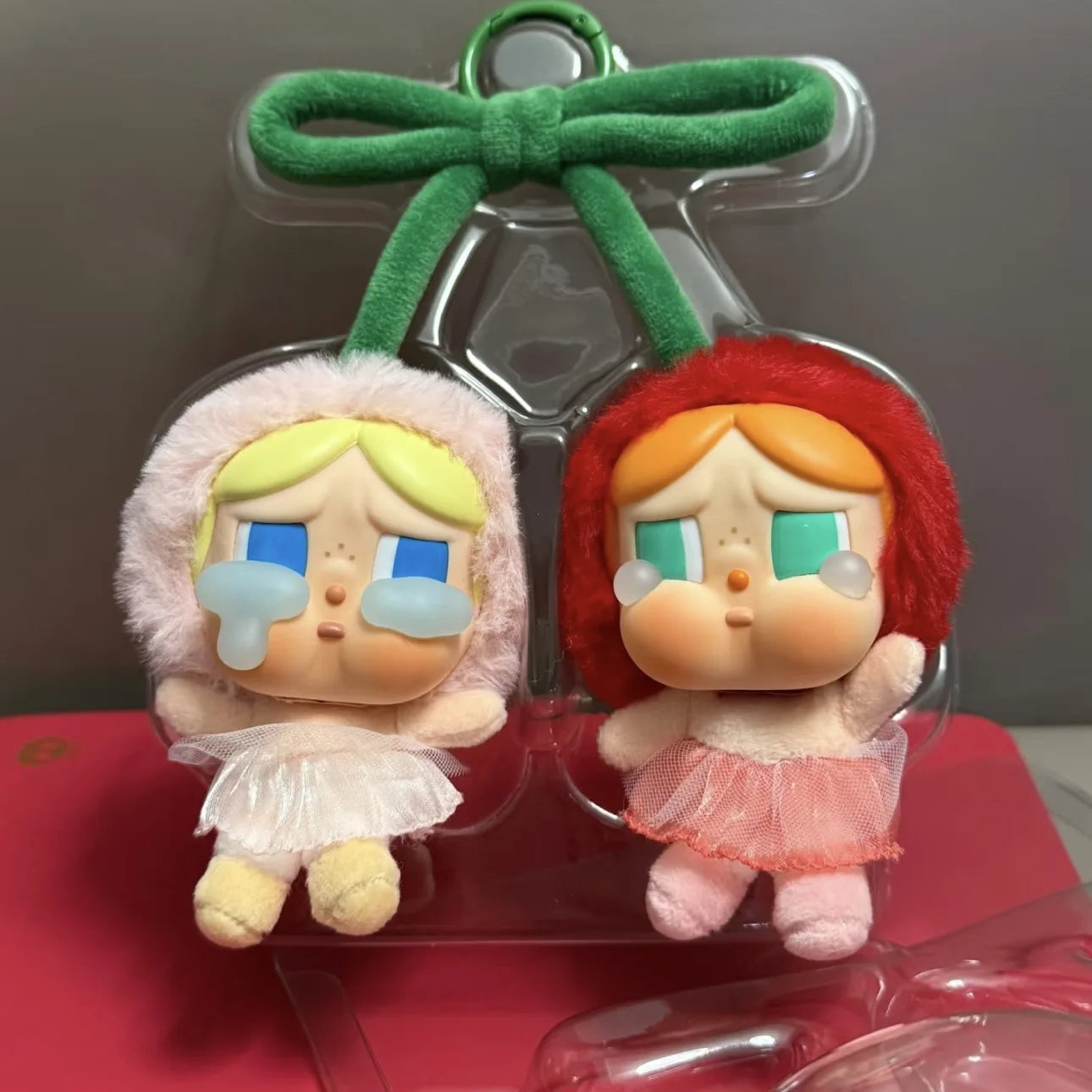 Pop Mart Cry baby, love you cherry much. Two small figurines with green hair bows in a clear plastic packaging on a red surface.