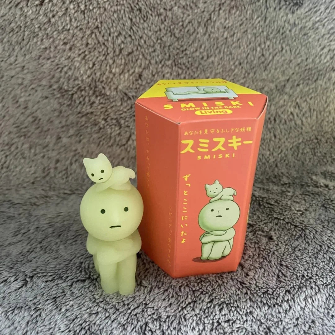 Smiski, living room series. Glowing green figurine of a character with a smaller figure on its head, next to its packaging box on a textured gray surface.