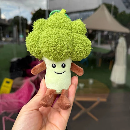 Jellycat. Plush toy shaped like a broccoli with a smiling face held in a hand outdoors.