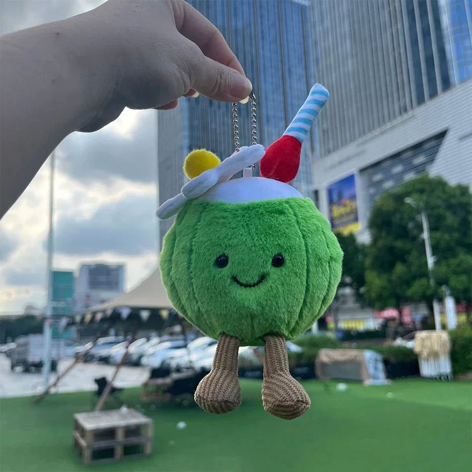 Jellycat Coconut. Plush toy resembling a green character with a straw and cherry on top, held by a hand outdoors.