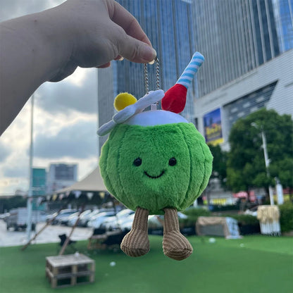 Jellycat Coconut. Plush toy resembling a green character with a straw and cherry on top, held by a hand outdoors.