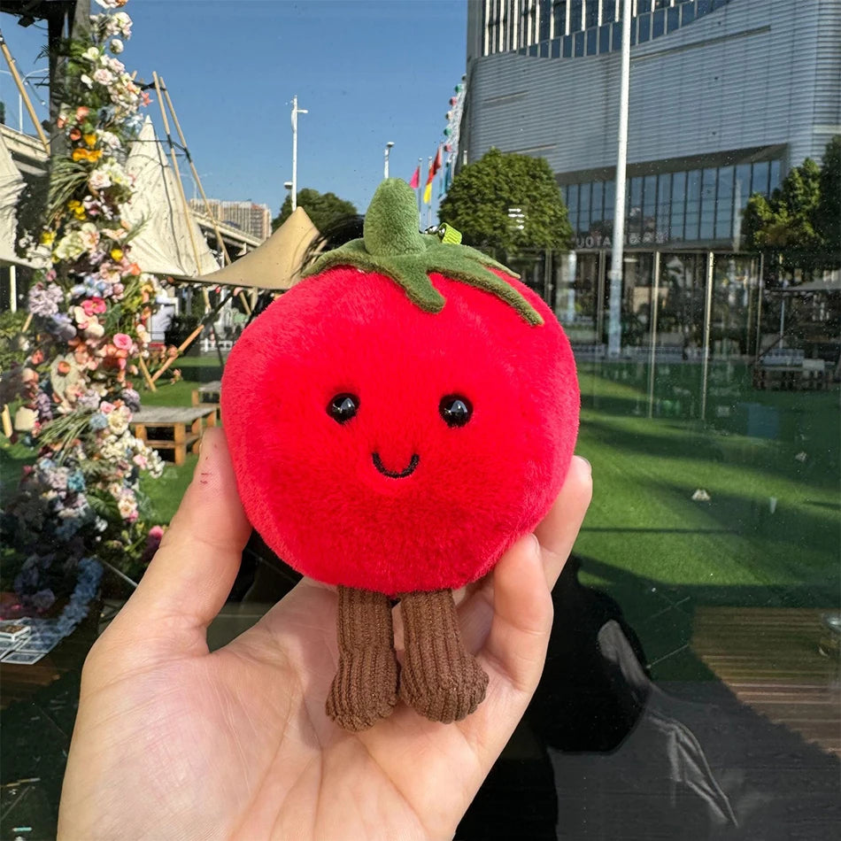 Jellycate. Plush tomato toy held in front of a cityscape with flowers and a building.