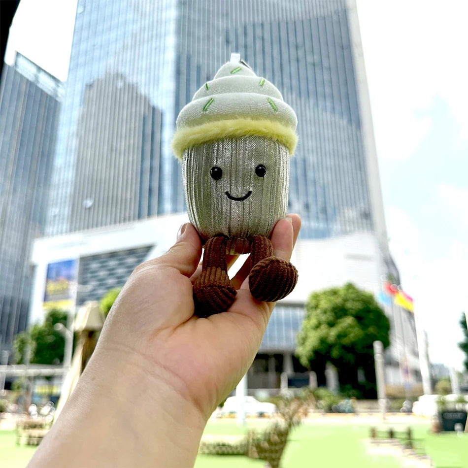 Jellycat. Toy acorn with a smiling face held in front of a cityscape