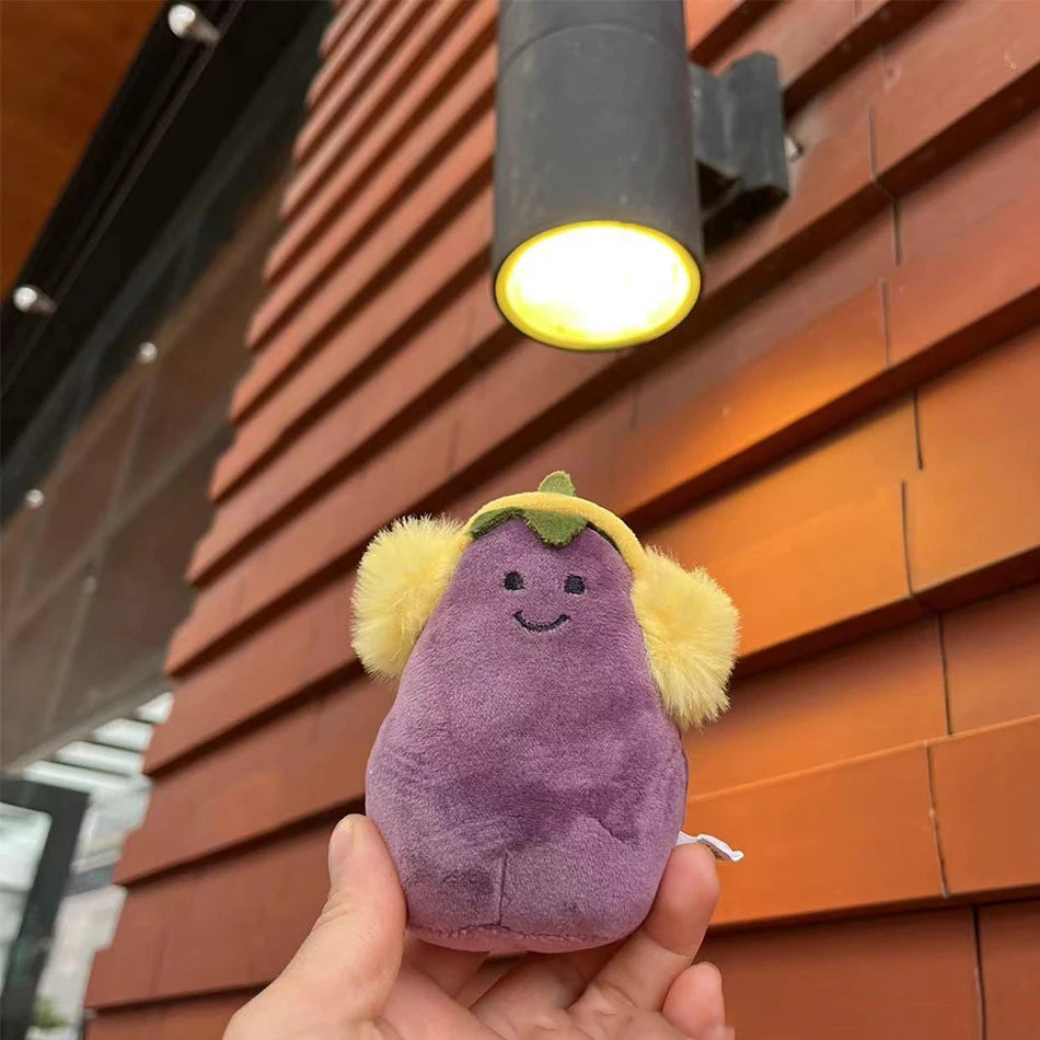 Jellycat. Plush toy resembling an eggplant with yellow earmuffs held in front of a building.