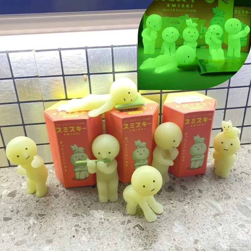 Smiski, living room series. Glowing yellow figures in front of red boxes with green packaging in the background.