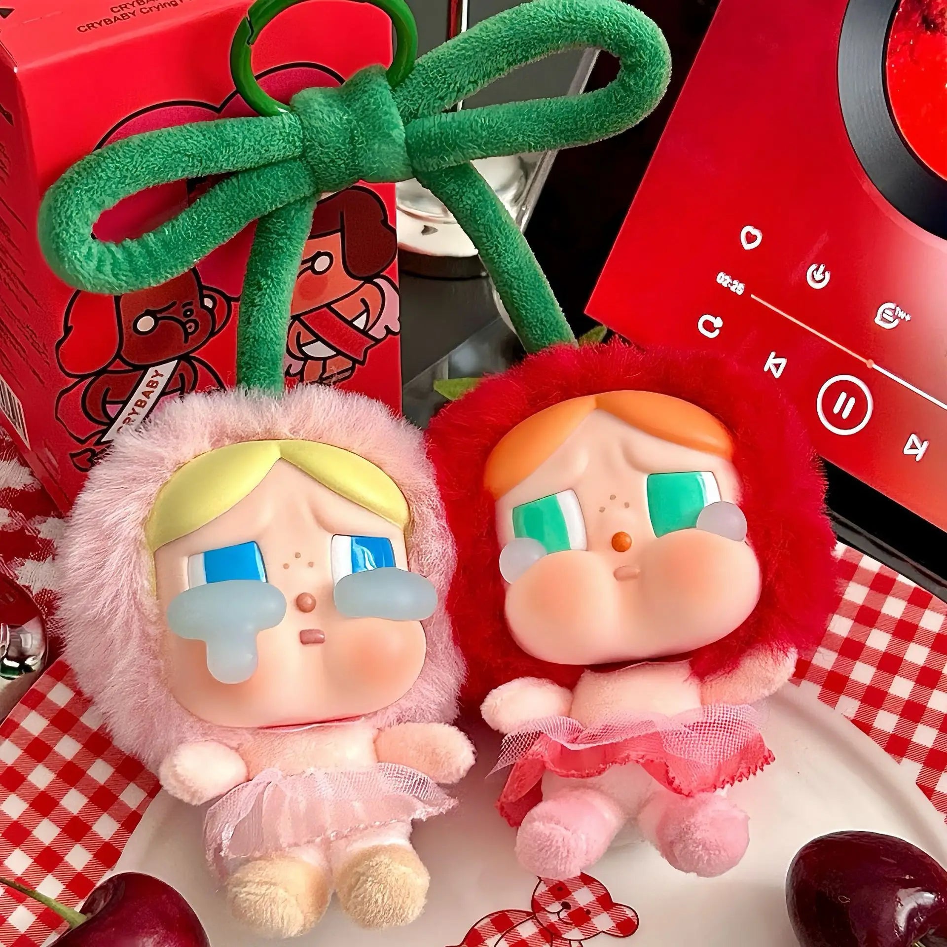 Pop Mart Cry baby, love you cherry much. Two plush toys with green bows on a checkered tablecloth with a red electronic device in the background.