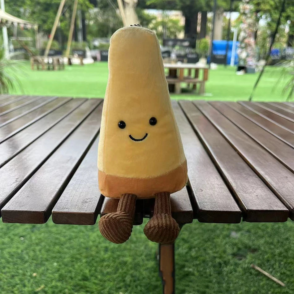 Jellycat Parmesan Cheese. Plush toy resembling a yellow ice cream cone with a smiley face on a wooden table outdoors.