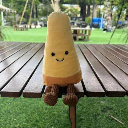 Jellycat Parmesan Cheese. Plush toy resembling a yellow ice cream cone with a smiley face on a wooden table outdoors.