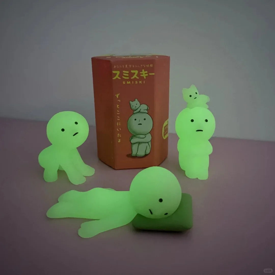 Smiski, living room series. Glowing green cartoon-like figures in front of a book with Japanese text on a gray background