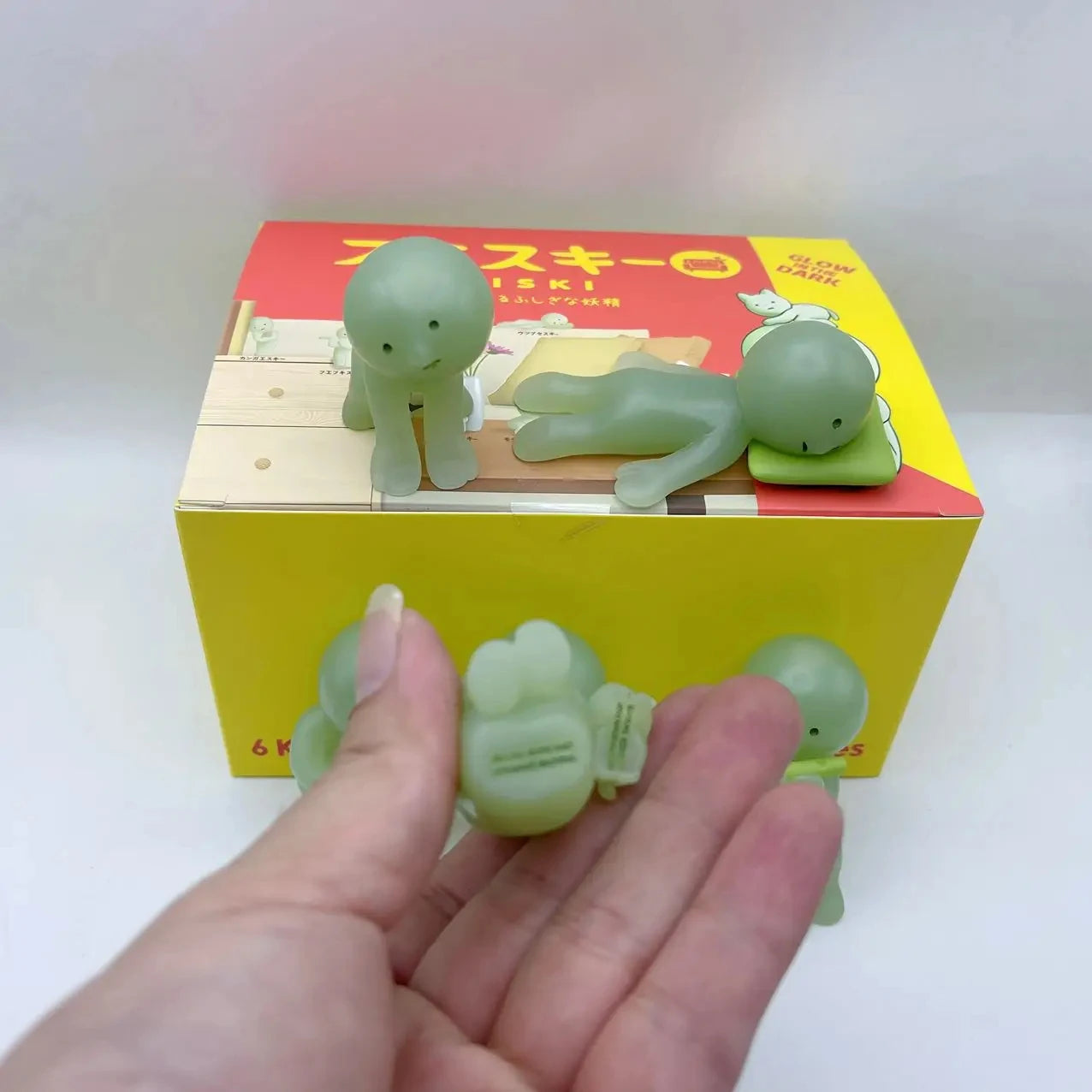Smiski, living room series. Green dinosaur-shaped erasers in front of a yellow box with Japanese text