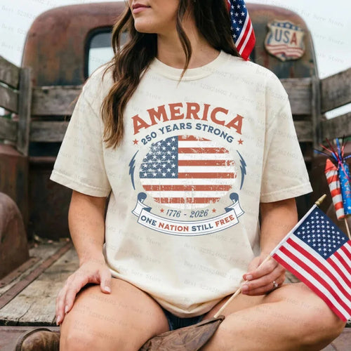 Person wearing a 'America 250 Years Strong' t-shirt with an American flag design, sitting in front of a vintage truck.