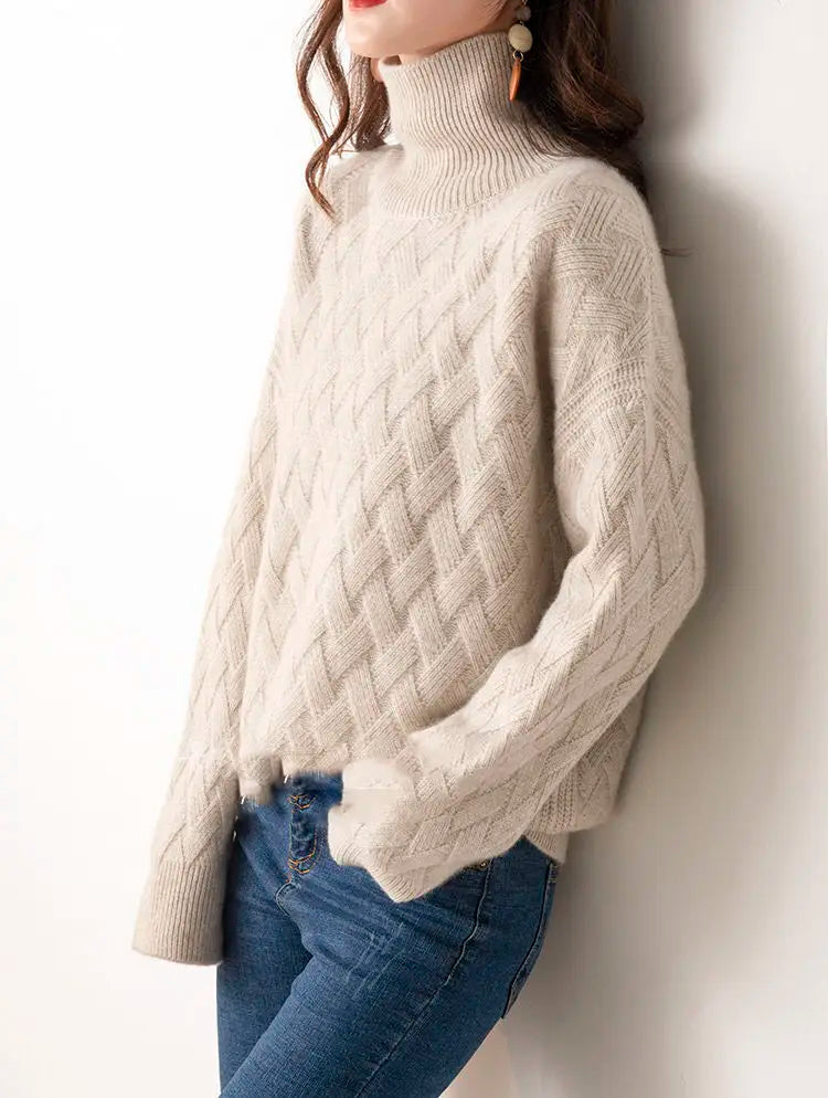 Person wearing a beige cable knit sweater and blue jeans against a white wall