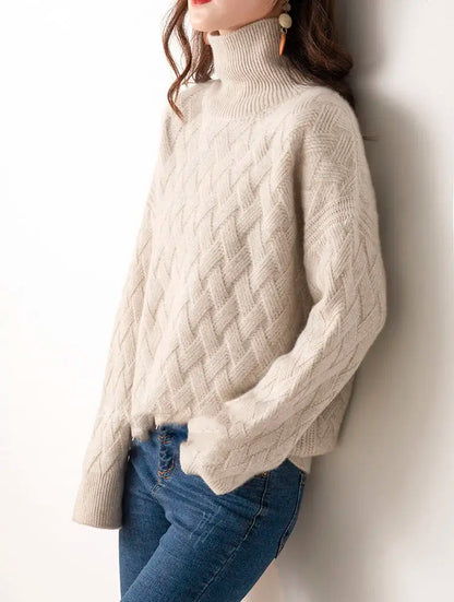 Person wearing a beige cable knit sweater and blue jeans against a white wall