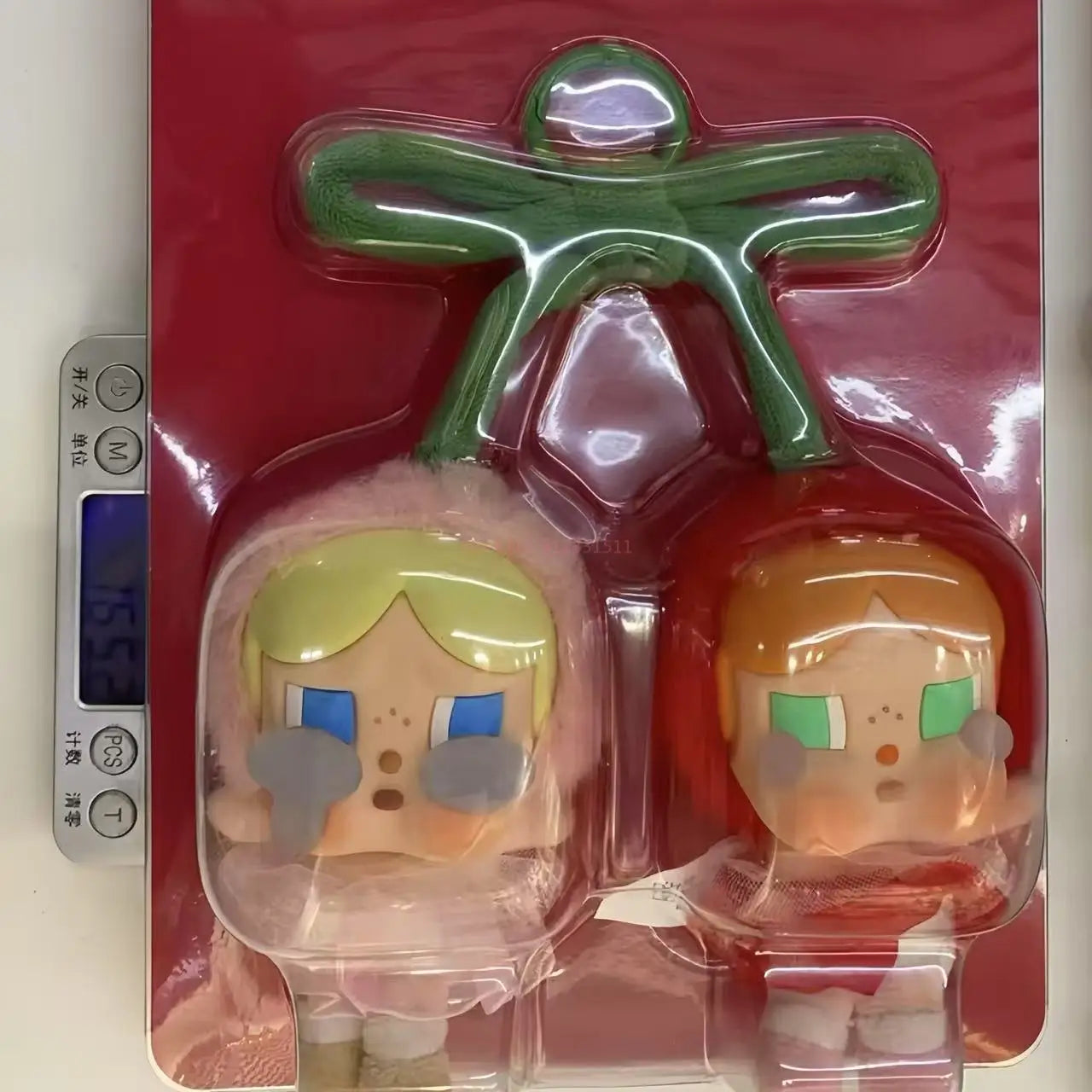 Pop Mart Cry baby, love you cherry much. Two cartoon character figurines in clear packaging on a digital scale with a red background.